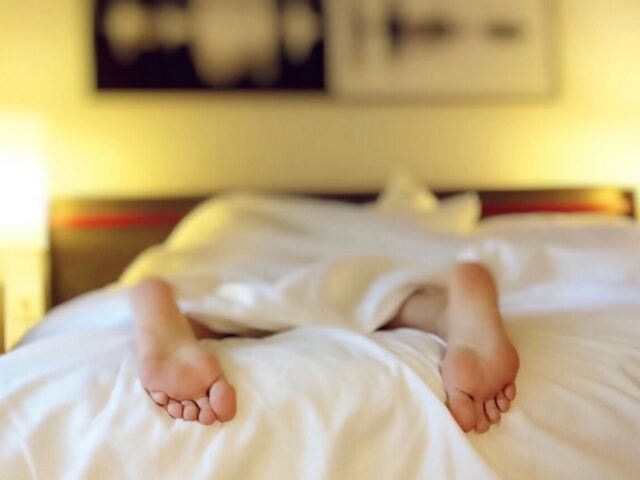 A person lying in bed with feet exposed under a white blanket, softly lit bedroom with warm lighting and abstract wall art.