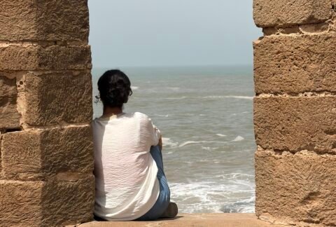 Woman sitting alone by the sea, looking towards the horizon through an opening in an old stone wall.